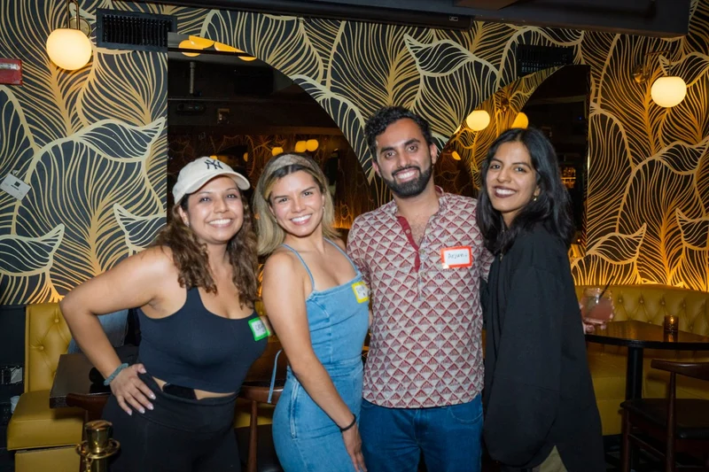 Four people smiling together at a bar with patterned wallpaper, warm lamps, and event name tags.